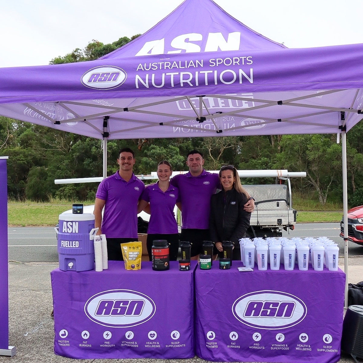 Four people standing behind an ASN Australian Sports Nutrition pop-up canopy and table setup outdoors. The setup includes branded purple canopy, matching purple table covers, a chilled drink dispenser, supplement containers, and a row of white shaker bottles.